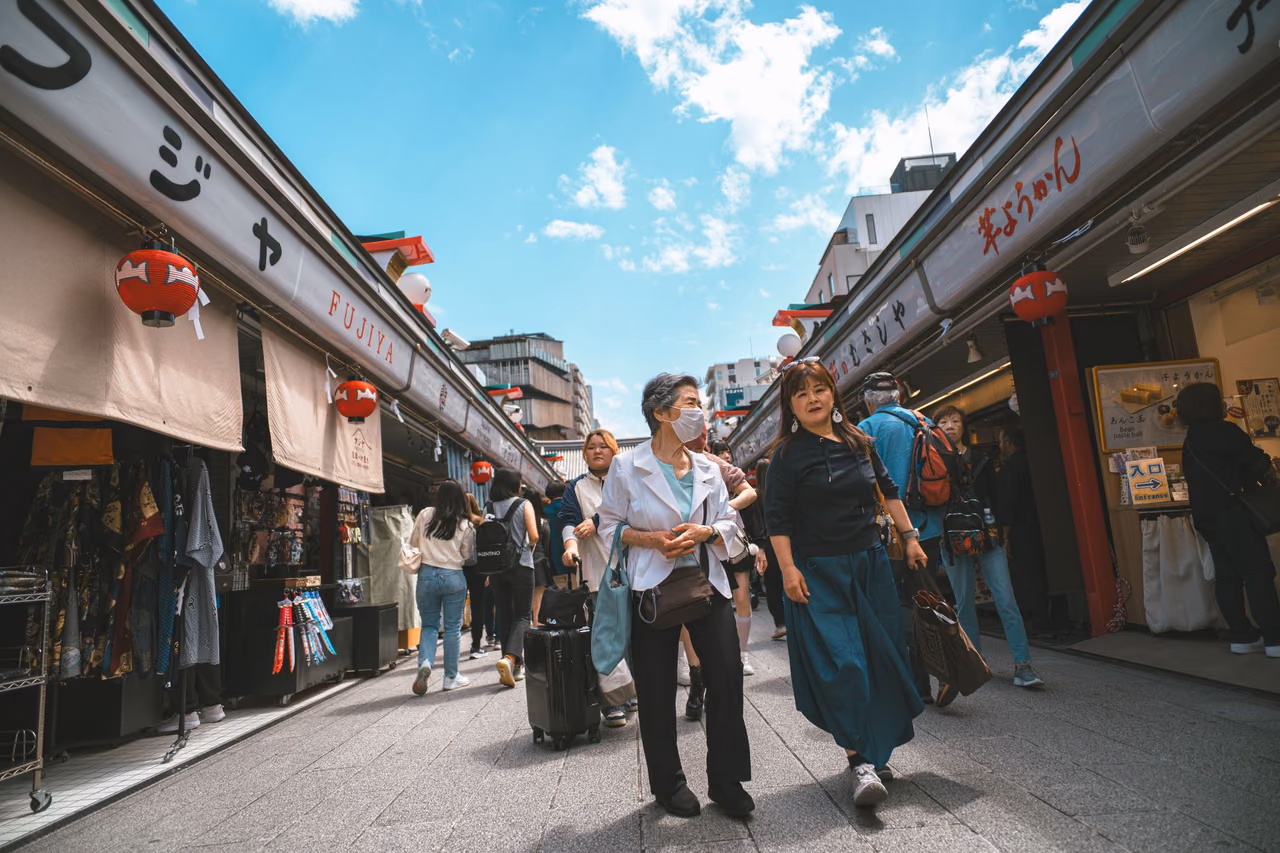Shoppers walk down a Tokyo shopping street lined with shops and red lanterns, one pulling a rolling suitcase under a blue sky