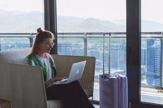A woman works on a laptop from a sofa by tall windows overlooking a city and mountains, with a rolling suitcase standing beside her.