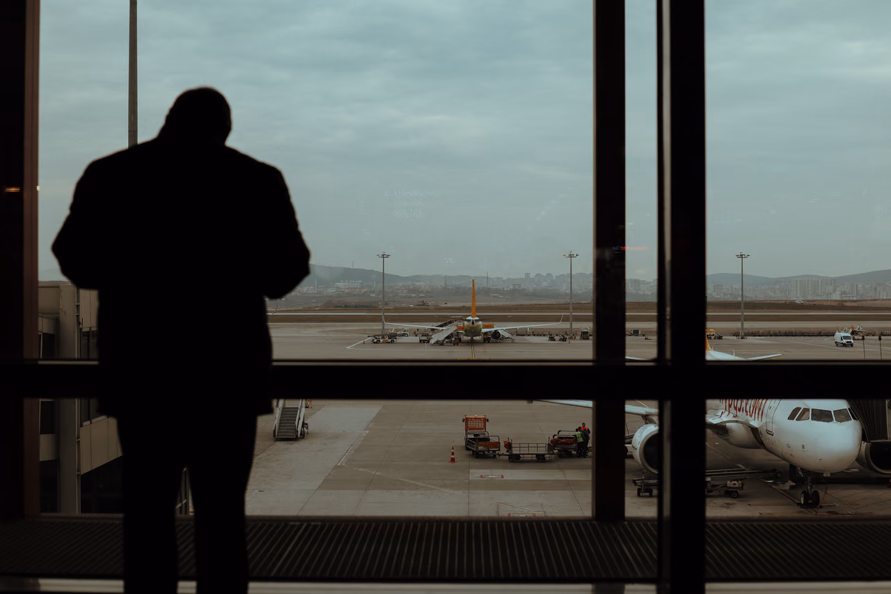 A traveler stands in silhouette at an airport terminal window looking out at parked jets on the apron.