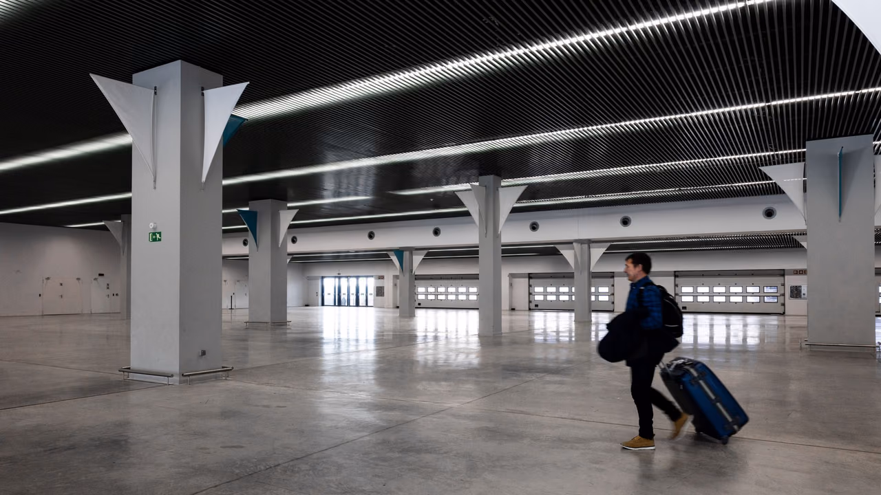 A traveler in a dark jacket pulls a blue rolling suitcase through a wide, nearly empty airport hall with white columns and long strips of ceiling lights.