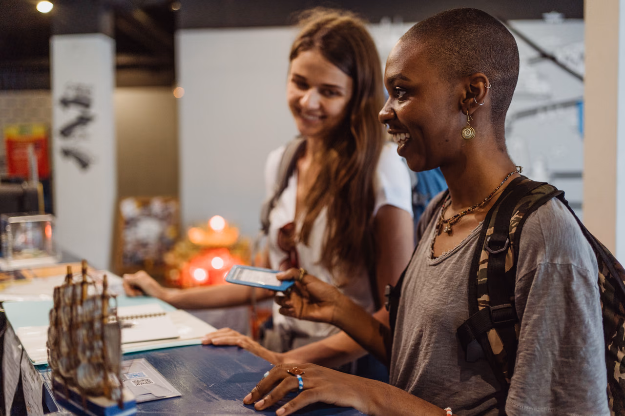 Two smiling travelers with backpacks checking in at a hotel reception desk, one holding a phone