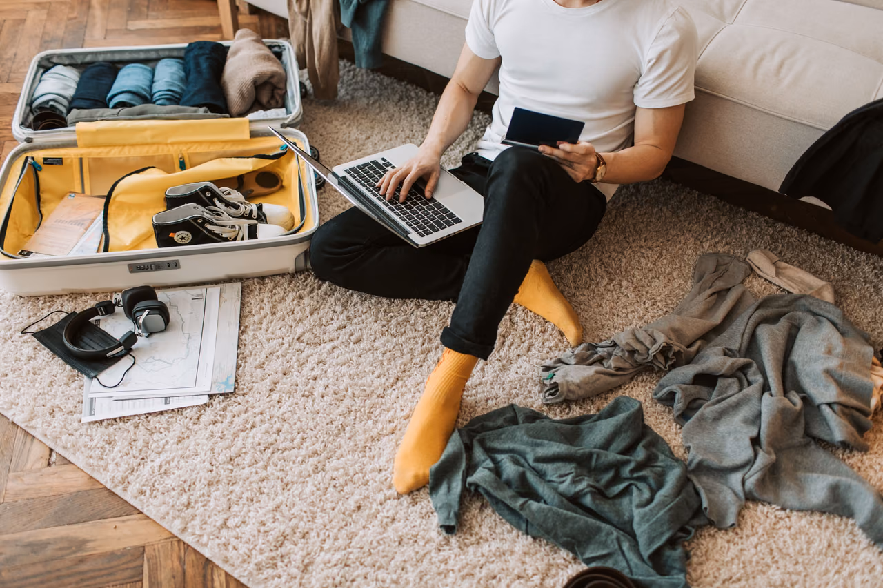 A man sits on a rug holding a passport and typing on a laptop next to an open, half-packed suitcase.