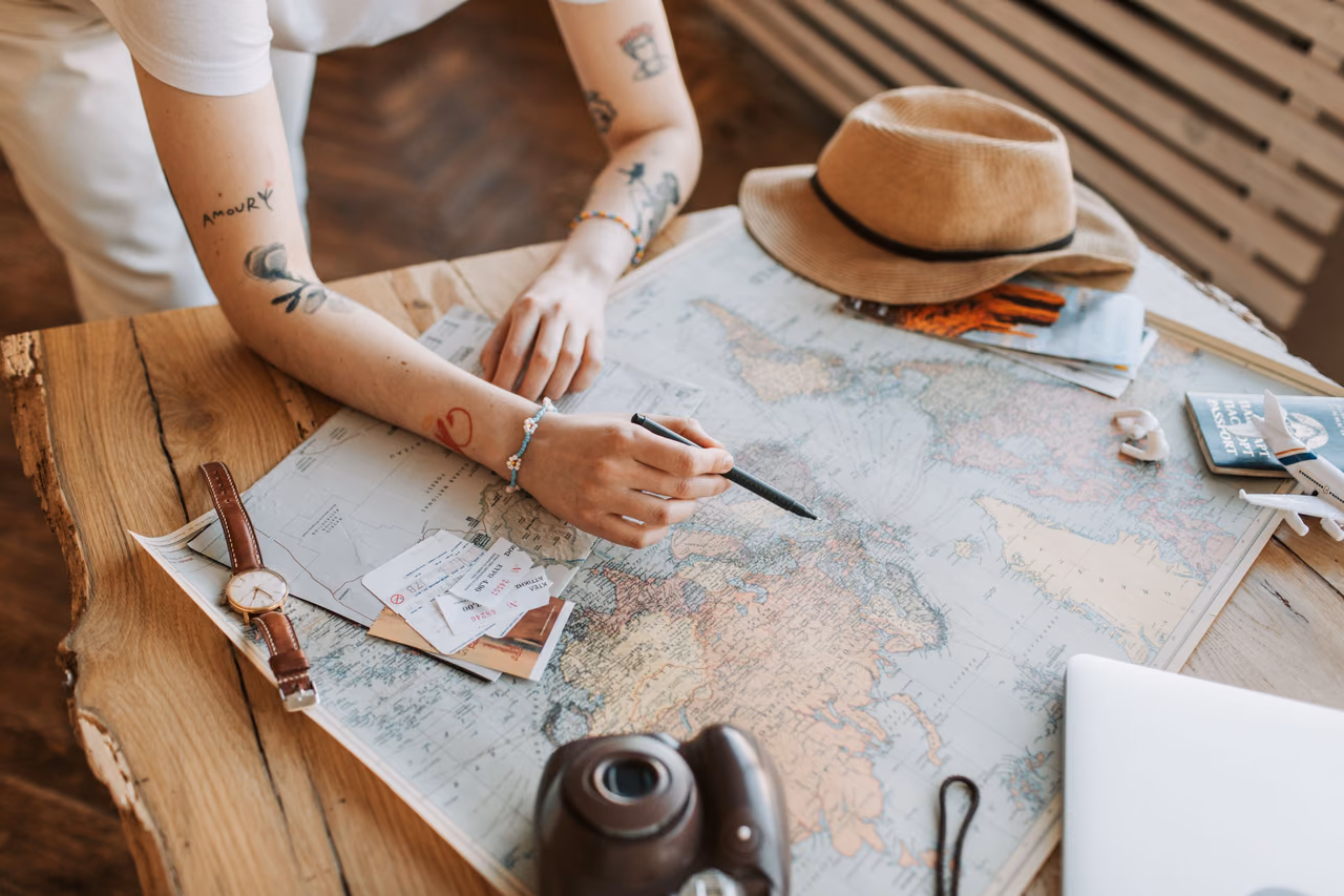 A traveler's hands marking a route on a world map spread across a wooden table, with tickets, a watch, a camera and a straw hat nearby.