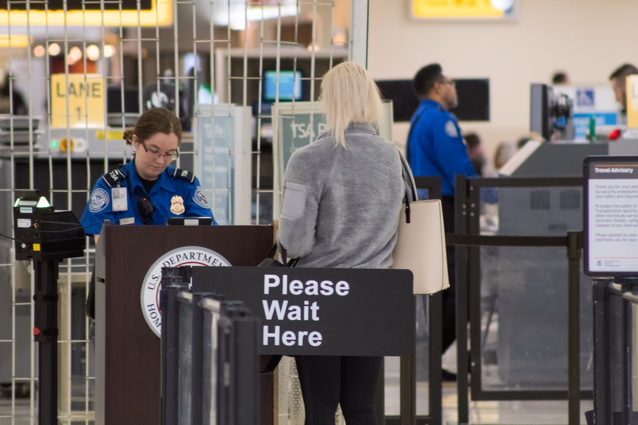 A TSA officer checks a traveler's documents at a podium behind a Please Wait Here sign at an airport checkpoint