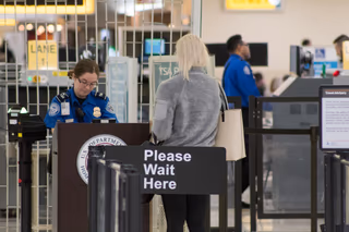 A TSA officer checks a traveler's documents at a podium behind a Please Wait Here sign at an airport checkpoint