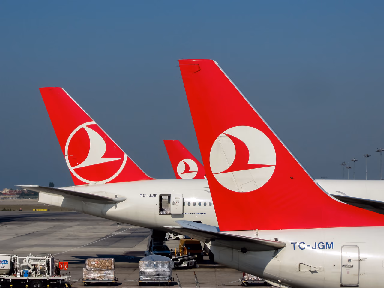 Red tail fins with the crescent logo on three Turkish Airlines jets parked on the ramp under a clear sky