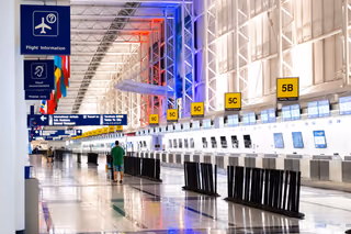 A long, mostly empty airport check-in hall with yellow zone signs, rows of self-service kiosks and stanchions on a polished floor