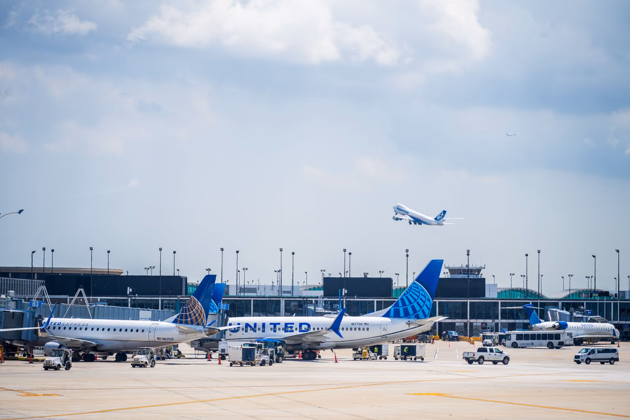 United Airlines jets parked at terminal gates under a cloudy sky while another jet climbs overhead after takeoff