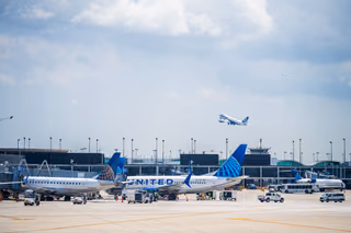 United Airlines jets parked at terminal gates under a cloudy sky while another jet climbs overhead after takeoff