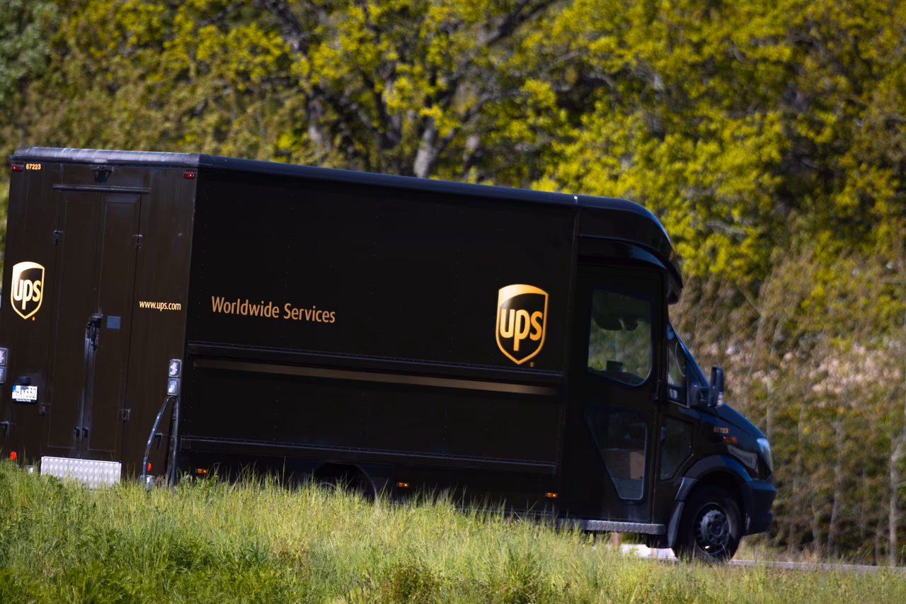 A brown UPS package delivery truck with the gold UPS shield on its side, driving past green trees and tall grass