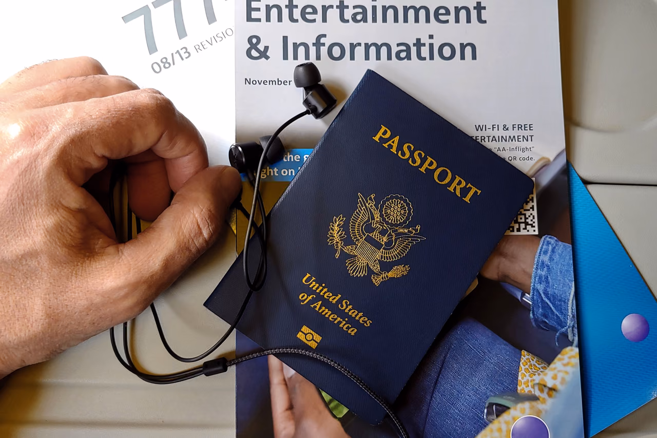 A navy U.S. passport resting on an in-flight magazine next to black earbuds on an airplane seat, with a traveler's hand beside it