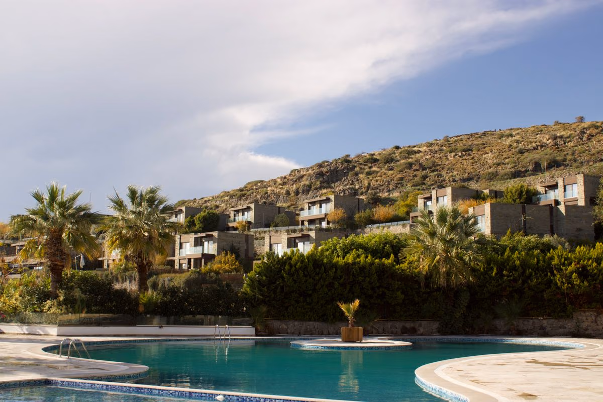 Stone vacation villas on a hillside behind a curved pool with palm trees under a partly cloudy sky