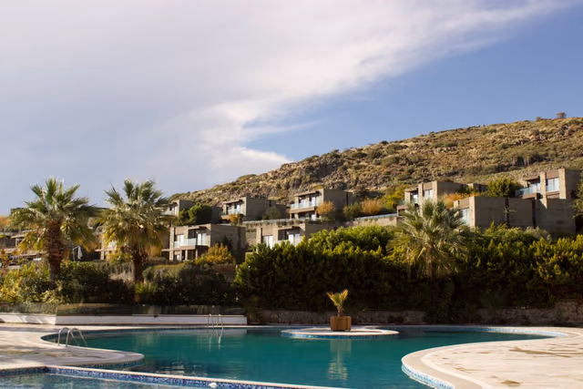 Stone vacation villas on a hillside behind a curved pool with palm trees under a partly cloudy sky