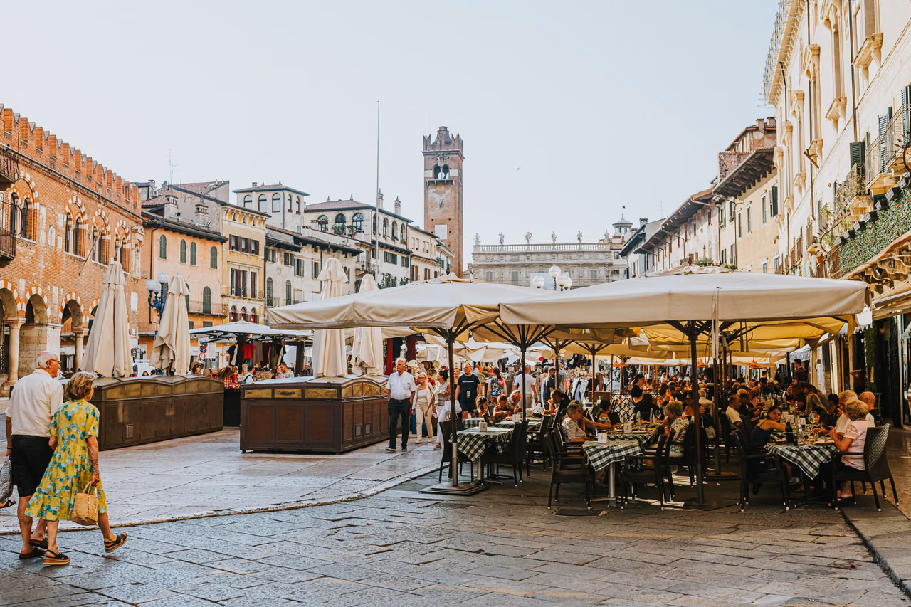 Outdoor cafe tables under large umbrellas on Piazza delle Erbe in Verona, Italy, with people dining and walking past historic buildings.