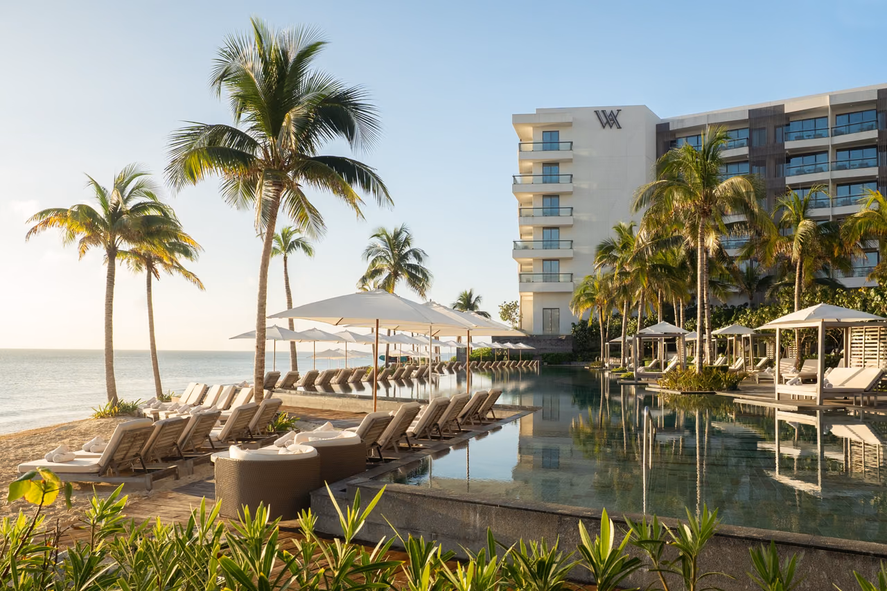 Infinity pool lined with white loungers and umbrellas beside the beach at Waldorf Astoria Riviera Maya, with palm trees and the resort's hotel building behind