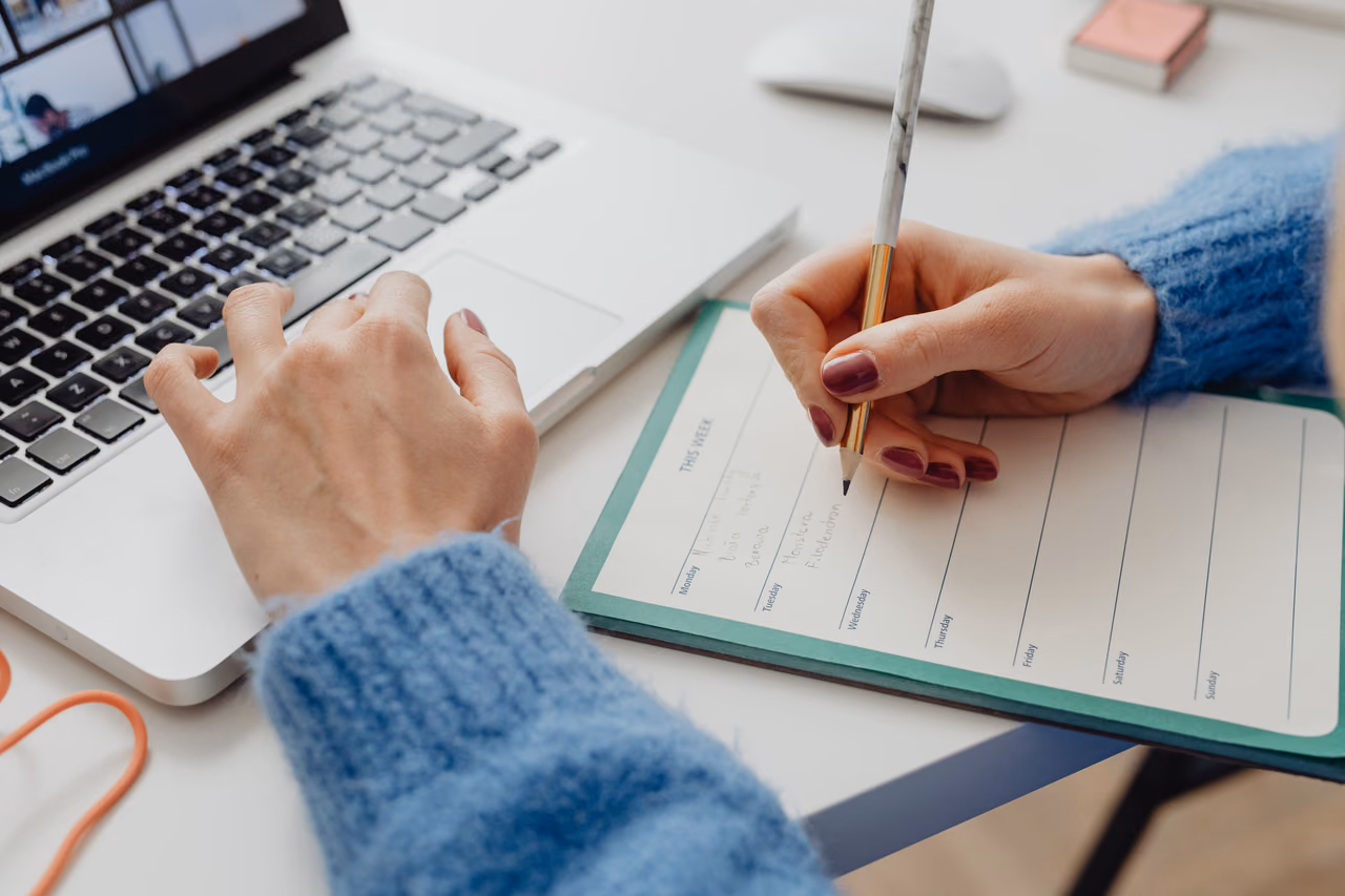A person in a blue sweater types on a laptop with one hand while filling in a weekly planner with the other.