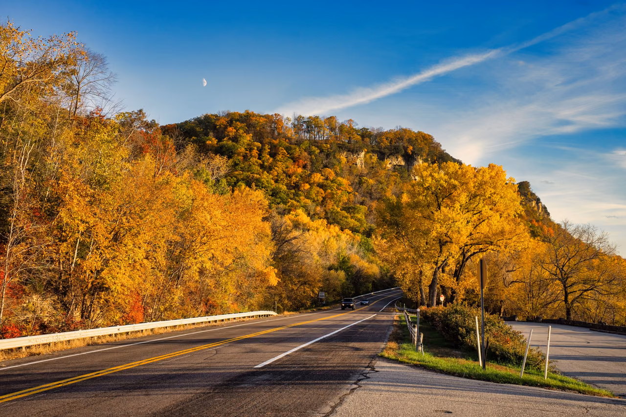 A two-lane highway curving past wooded bluffs in gold and orange fall foliage under a blue sky